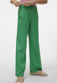 Zielone, szerokie spodnie wykonane z gładkiego materiału, z elastycznym paskiem w talii i falbaniastym wykończeniem. Połączone z lekkimi, plecionymi butami.