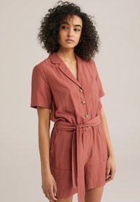 Kortärmad jumpsuit i rostfärgad nyans, tillverkad av ett lätt material, med krage, knappslå framtill och knytbart midjebälte samt sidofickor.