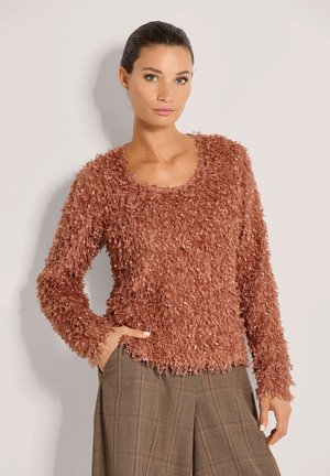 Vrouw met donker haar in een getextureerde, pluizige roestkleurige trui en bruine geruite broek, staand met één hand in haar zak tegen een effen achtergrond.