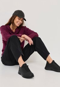 Melni sporta apavi ar teksturētām virsmām, apvienoti ar melnām platām biksēm un bordo garām piedurknēm kreklu, nēsāti ar melnu cepuri.