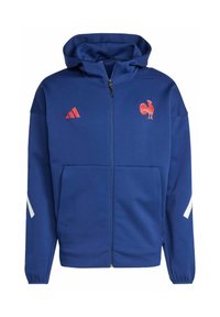 Sudadera azul con cremallera hecha de un tejido suave, que presenta un logotipo de gallo rojo y rayas rojas de Adidas en el pecho, además de acentos blancos en las mangas.