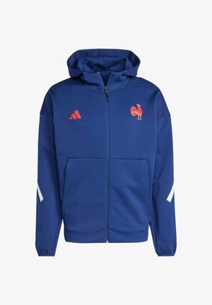 Sudadera azul con cremallera hecha de un tejido suave, que presenta un logotipo de gallo rojo y rayas rojas de Adidas en el pecho, además de acentos blancos en las mangas.