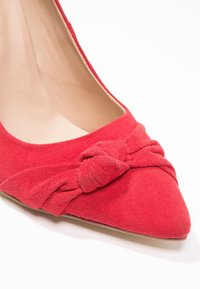 Chaussure à talon haut en daim rouge avec un bout pointu et un détail noué à l'avant. Doublure intérieure lisse ; talon fin et délicat.