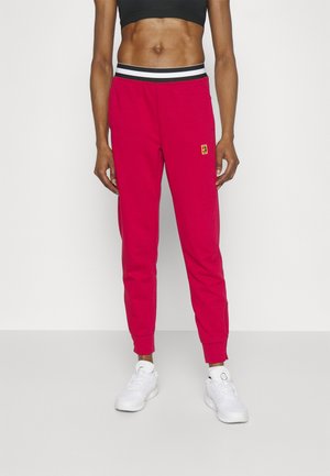 Fit osoba w jasnoczerwonych spodniach dresowych z paskiem w paski, czarnym krótkim topie i białych butach sportowych na prostym tle.