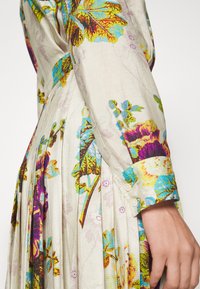 Šaty s květinovým vzorem v živých odstínech zelené, modré a fialové. Vyrobeny z texturované tkaniny s záhyby a detaily na manžetách s knoflíky.