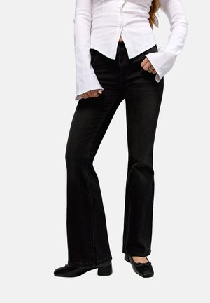 Woman wearing black flared jeans, a white fitted shirt with flared cuffs, black low-heeled shoes, and rings on both hands.