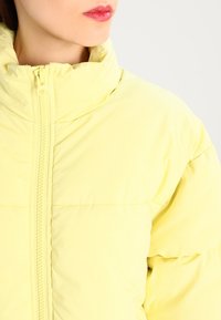 Chaqueta acolchada amarilla con cuello alto, que cuenta con una cremallera central y un diseño acolchado. Textura suave y material ligero.