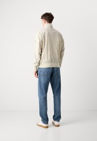 Lys beige bomberjakke med elastiske mansjetter og søm, parret med blå jeans og hvite joggesko. Har et minimalistisk design.
