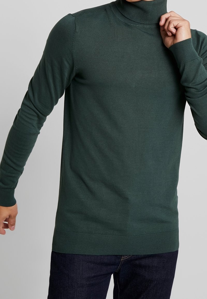 Dunkelgrüner Rollkragenpullover aus glattem Strickstoff; taillierte Passform mit langen Ärmeln und gerippten Bündchen und Saum. Trägt er zu dunklen Jeans.