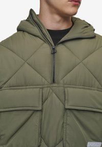 Giacca a modo con cappuccio di colore verde oliva, con un design trapuntato, chiusura con zip e grandi tasche a toppa. Presenta una superficie liscia e imbottita.