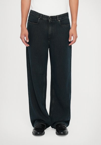 Una persona che indossa jeans di denim neri larghi e scarpe di pelle nera, con le mani rilassate sui fianchi su uno sfondo chiaro e uniforme.