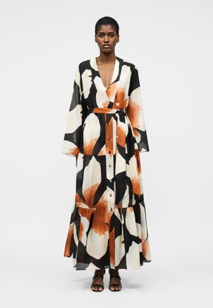 Γυναίκα που φοράει ένα μακρύ, κουμπωτό φλοράλ φόρεμα με μαύρα, λευκά και πορτοκαλί πέταλα, δεμένο με ζώνη που ταιριάζει, στέκεται μπροστά σε ένα μονόχρωμο φόντο.