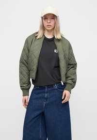 Νέα γυναίκα που φοράει εκρού καπέλο baseball, πράσινο πάλλευκο μπουφάν bomber, μαύρο μπλουζάκι και φαρδιά τζιν μπλε απέναντι σε απλό φόντο.