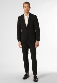 Zwart pak met een getailleerd ontwerp, bijpassende zwarte broek en een lichtgekleurde shirt. Leren schoenen maken de formele uitstraling compleet.