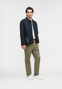 Veste légère marine portée sur un pull crème, associée à un pantalon vert olive et des baskets blanches. Sac bandoulière noir suspendu à l'épaule.
