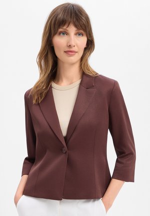 Vrouw met een lichte huid en schouderlang bruin haar, die een kastanjebruine blazer draagt over een beige top, met haar handen in de zakken van een witte broek.