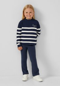 Maglione a righe blu navy e bianco con scollatura rotonda e dettaglio a fiocco argento sul petto. Abbinato a leggings blu navy e scarpe da ginnastica bianche.