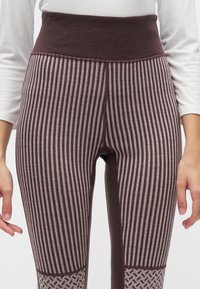 Κοντινό πλάνο ενός ατόμου που φοράει leggings με ψηλή μέση και κάθετες καφέ και μπεζ ρίγες, με διακοσμητικές θήκες στα γόνατα, συνδυασμένα με λευκό μακρυμάνικο τοπ.