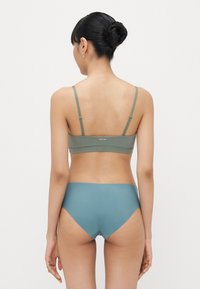 Grüne Bralette mit verstellbaren Trägern, kombiniert mit hochgeschnittenen blauen Unterhosen. Glatte Textur und minimalistisches Design von hinten sichtbar.