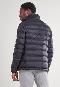 Giacca piumino blu navy con vestibilità slim, trapuntatura orizzontale, colletto alto e una texture liscia e lucida. Presenta un logo laterale sul petto.