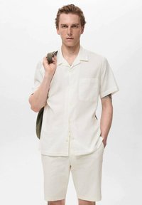 Cămașă cu mânecă scurtă, de culoare bej deschis, cu nasturi și o buzunare, asortată cu pantaloni scurți bej; textură netedă, croială lejeră.