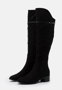 Botas negras por encima de la rodilla hechas de suave ante. Tienen un ligero tacón, punta afilada y una correa decorativa negra cerca de la parte superior. Superficie texturizada.