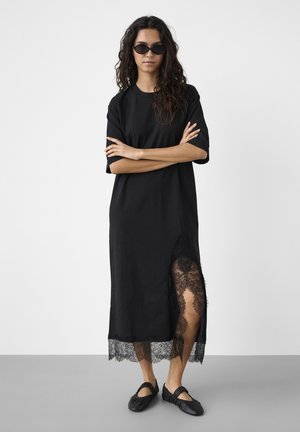 Kvinde med langt, krøllet hår iført sorte solbriller, en sort kjole med blonder i siden og sorte flade sko, stående med armene krydset.