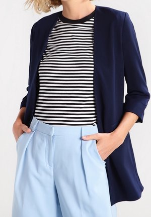 Femme portant un blazer bleu marine sur une chemise rayée noire et blanche avec un pantalon taille haute bleu clair, mains dans les poches.