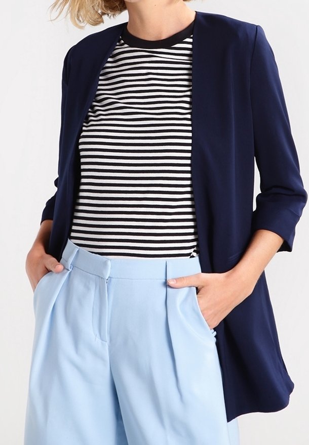 Femme portant un blazer bleu marine sur une chemise rayée noire et blanche avec un pantalon taille haute bleu clair, mains dans les poches.