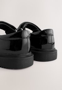 Paar glänzende schwarze Lederschuhe mit dicken schwarzen Sohlen, von hinten auf einem schlichten hellen Hintergrund gesehen.