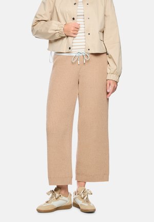 Lower body of a person wearing beige ribbed knit wide-leg pants, beige and white sneakers with satin laces, and a beige jacket over striped top.