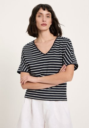 Frau mit schulterlangem dunklem Haar, trägt ein schwarz-weiß gestreiftes V-Ausschnitt-Shirt und weiße Hose, die Arme verschränkt.