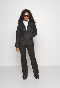 Black puffer jacket with quilted design and orange accents, worn over a light-colored turtleneck, paired with dark pants and ski boots.