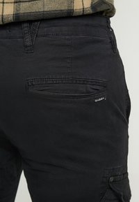 Närbild av svarta byxor med bakficka och liten "GABBA"-märkesetikett, bärs med en beige och svart rutad skjorta.