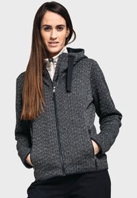 Chaqueta gris texturizada con cremallera frontal, capucha y bolsillos laterales. Presenta un patrón geométrico y cremallera negra contrastante con acentos.