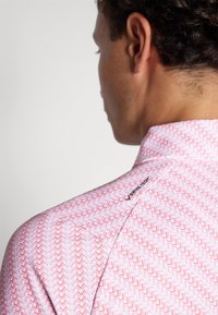 Short-sleeve shirt featuring a pink and white zigzag pattern, with a collar and a logo detail on the shoulder. Smooth texture.