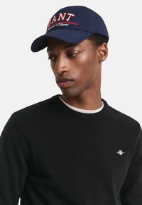 Cappellino da baseball blu con logo ricamato "GANT" rosso e bianco, abbinato a un maglione nero con un piccolo logo sul petto.