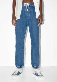 Persoon in blauwe spijker joggerbroeken met elastische boorden, witte en blauwe sneakers en een wit opengehaakt mouwloos topje.