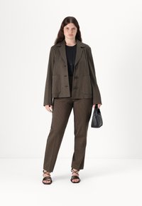 Blazer marron à motifs et pantalon assorti, tous deux en tissu texturé. Le mannequin tient un petit sac à main noir. Des talons noirs à lanières complètent le look.
