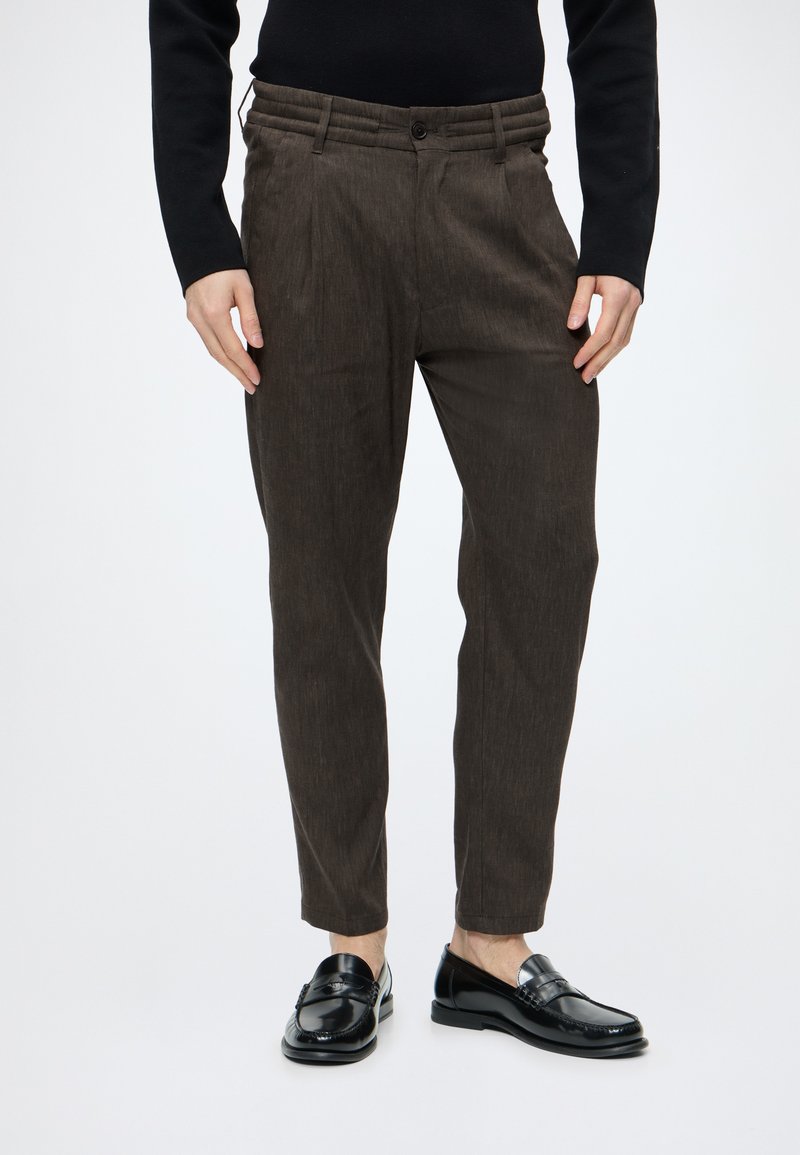 Homme portant un pantalon slim marron foncé, un haut noir à manches longues et des mocassins noirs brillants, debout devant un fond uni.