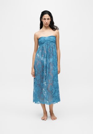 Vrouw staat blootsvoets in een strapless, blauwe doorschijnende kanten jurk met een bloemenpatroon tegen een effen witte achtergrond.