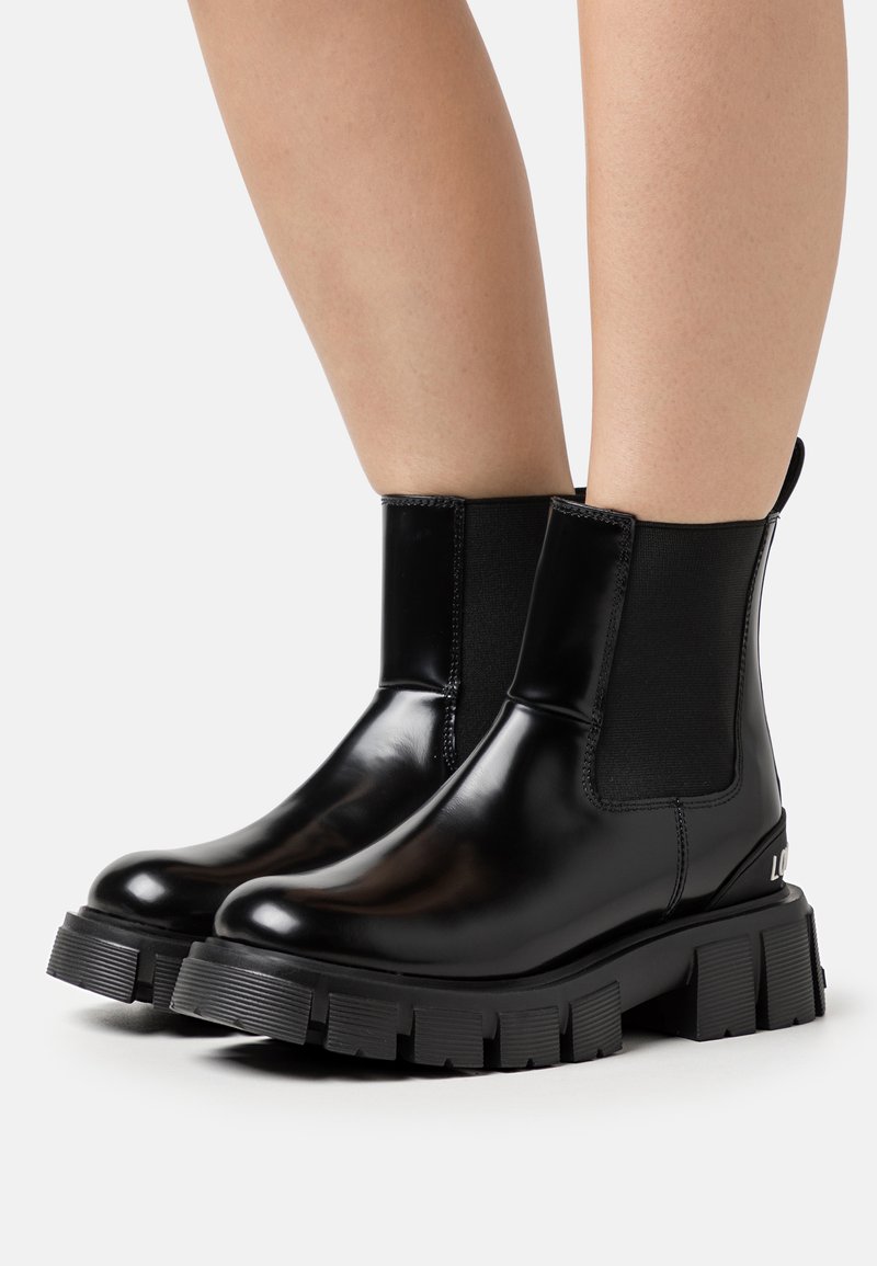 Botas de tornozelo pretas brilhantes com painéis laterais elásticos, sola texturizada espessa, e um detalhe com o logótipo. Acabamento suave com um design elegante.