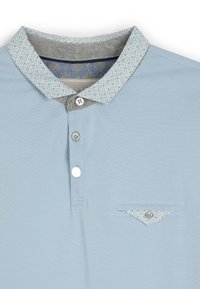 Jasnoniebieska koszulka polo z wzorzystym kołnierzykiem. Kołnierzyk ma szare i białe wzory geometryczne. Zawiera trzy guziki i kieszeń.