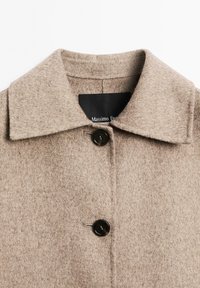 Beige uldfrakke med klassisk krave, enkelt sort knaplukning og en struktureret stoffinish. Mærke: Massimo Dutti.