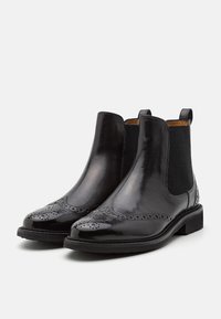 Bottes Chelsea en cuir noir avec des détails brogués, des panneaux latéraux élastiques, des languettes de tirage et des talons bas sur un fond clair uni.