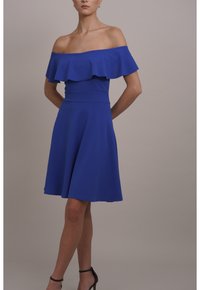 Kvinna som bär en kungligt blå off-shoulder klänning med volangdetaljer och svarta högklackade sandaler, stående mot en enkel bakgrund.