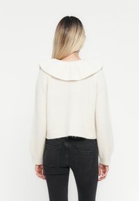 Kremfarget, kort genser med bred, brettet krage og ribbede mansjetter. Laget av mykt, teksturert materiale. Kombinert med mørkeblå jeans.
