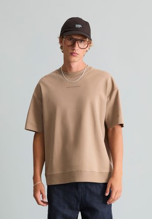 Jonge man draagt een beige oversized T-shirt, donkere jeans, zwarte pet, grote bril, zilveren ketting en armband, staat tegen een effen achtergrond.