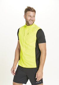 Neon-gelbe ärmellose Jacke mit hohem Kragen, Reißverschluss und reflektierenden Akzenten. Über einem schwarzen kurzärmeligen Hemd getragen.