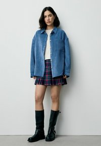Camicia azzurra in corduroy con bottoni, top bianco, mini gonna a quadretti in blu navy e rosso, stivali neri alti fino al ginocchio con dettaglio a fibbia.